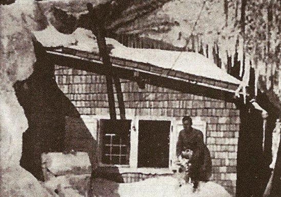 A person stands in front of a snow-covered cabin with icicles hanging above.