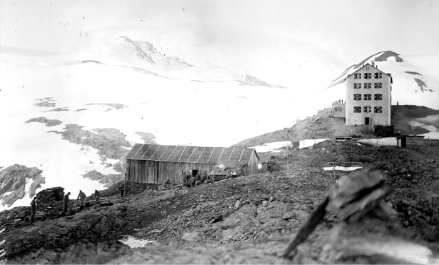 Historic mountain landscape featuring a building and lodge amidst snow-capped peaks.