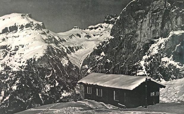 A rustic cabin surrounded by snow-capped mountains and rugged terrain.