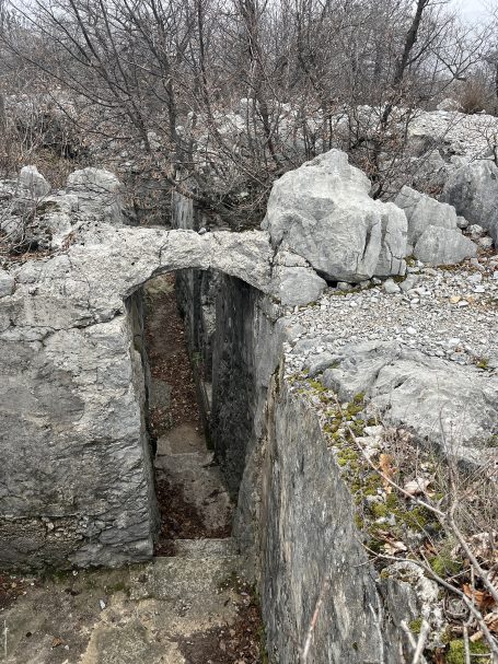 Ruins of stone structures with an archway amidst sparse trees and rocky terrain.