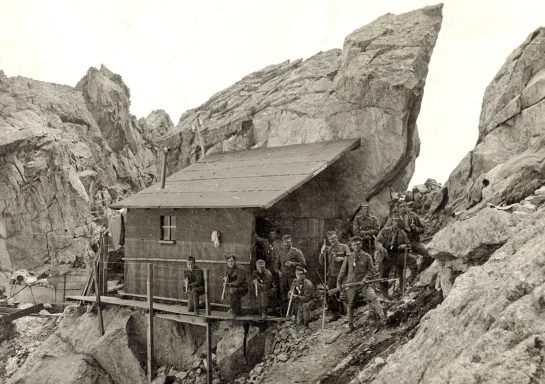 Historic wooden shack built against rocks, surrounded by soldiers on rugged terrain.