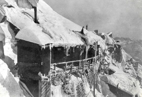 Old wooden cabin draped in snow and ice on a rocky mountainside.