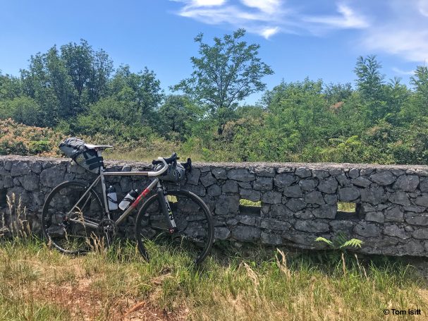 A black bicycle leaning against a stone wall with greenery in the background.