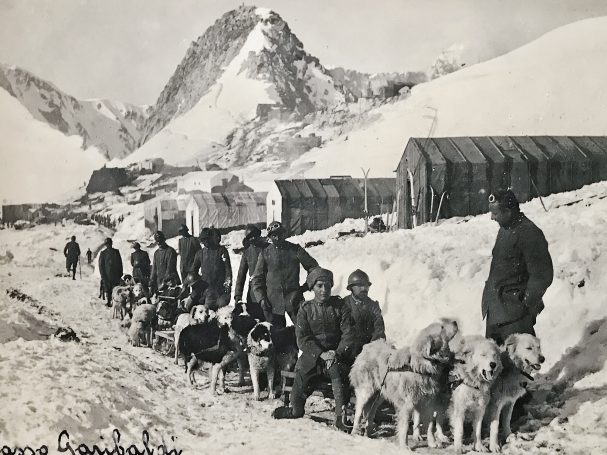 A line of people and sled dogs in snowy mountains near structures.