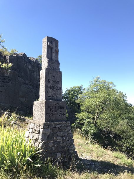 Stone monument with a tall, narrow structure set among greenery and clear blue sky.