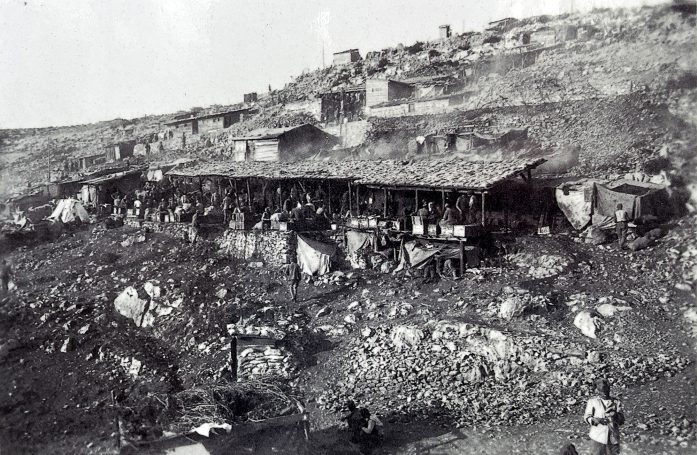 Historical black-and-white photo of a hillside settlement with makeshift buildings and people.