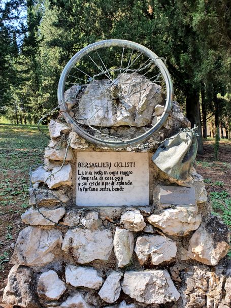 A stone monument featuring a bicycle wheel and a plaque in a wooded area.