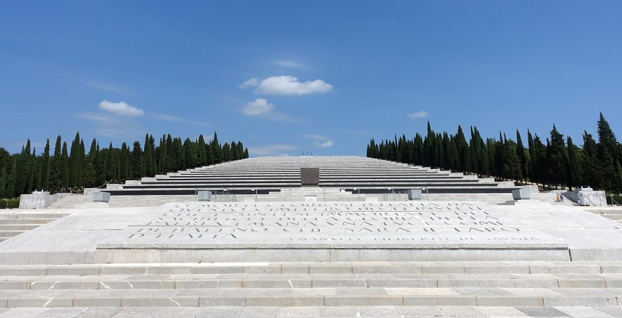 A large stone monument with steps leading up, surrounded by trees under a blue sky.
