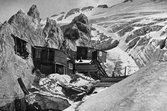 Two wooden huts near a snowy mountain landscape, surrounded by jagged ice formations.