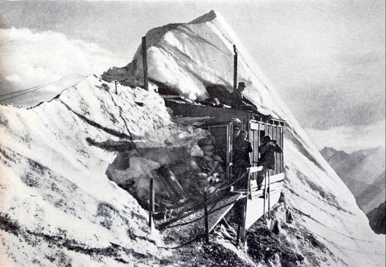 A historic mountain hut partially covered by snow and ice, surrounded by rugged peaks.