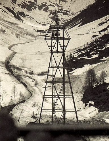 Black and white image of a tall tower beside a river in a mountainous landscape.