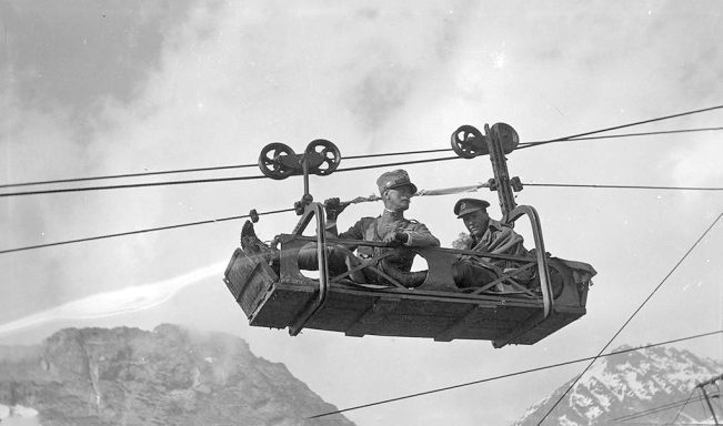 Two men sitting in a suspended lift, with mountains in the background.