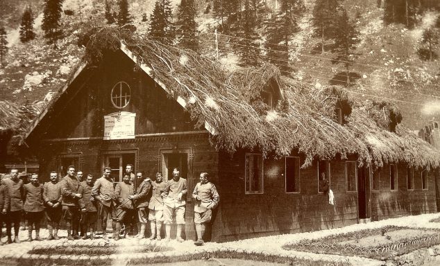 Group of soldiers in vintage uniform standing in front of a thatched-roof building.