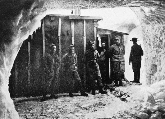 Soldiers standing at the entrance of a snow cave, with wooden structures in the background.