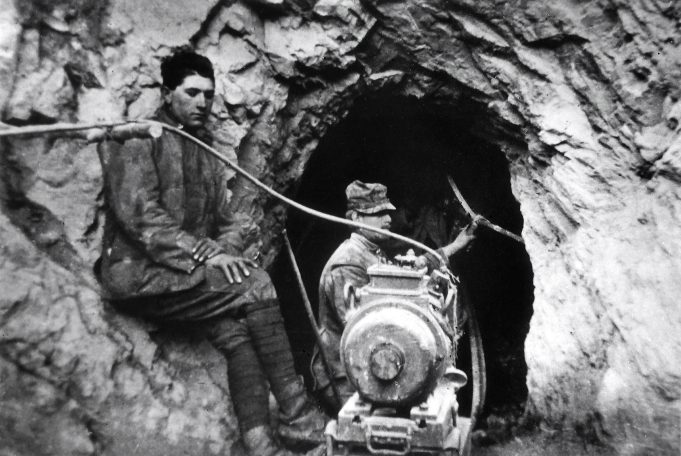 Two men near a mine entrance, one operating machinery, the other sitting beside him.