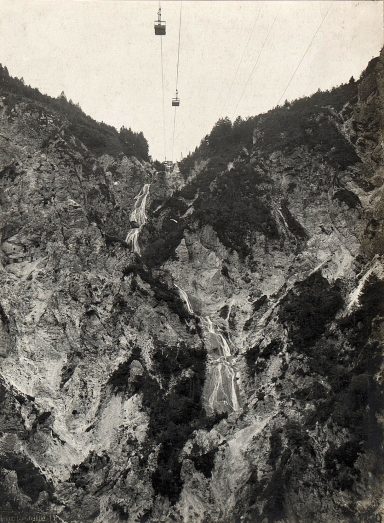 A cable car travels over a rugged mountain valley with steep cliffs.