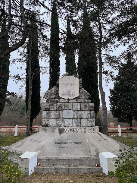 Stone monument surrounded by tall trees, with a plaque at the top and low white pillars.