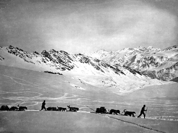 Two figures walking with sled dogs across a snowy landscape beneath mountains.