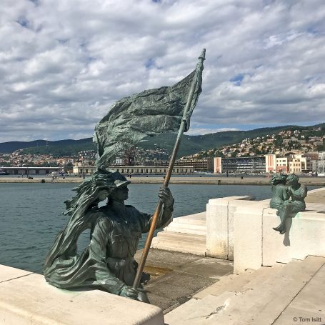 Bronze statues holding flags near water, with a cityscape and cloudy sky in the background.