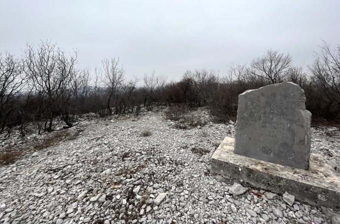 Rocky landscape with a prominent stone marker amid bare trees under a grey sky.
