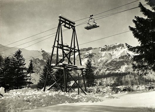 A wooden ski lift structure with a chair ascending over a snowy landscape and mountains.